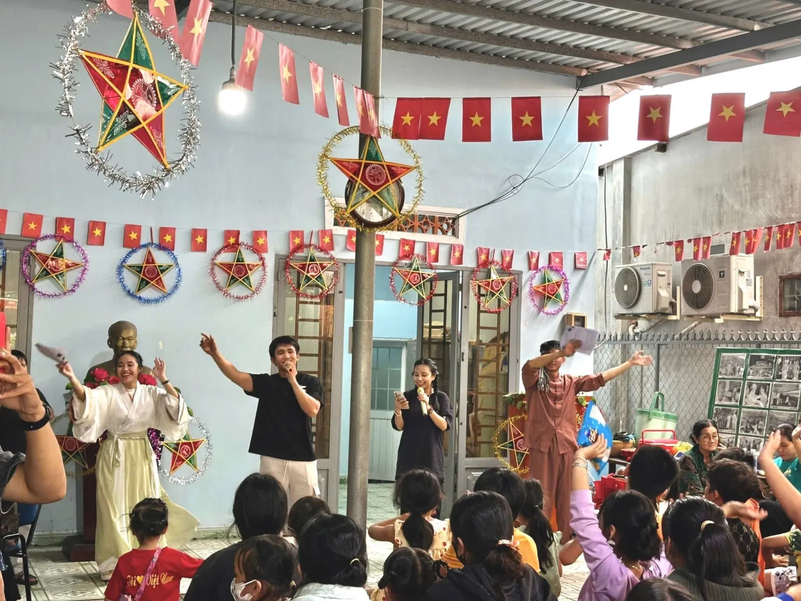 Các bạn tình nguyện viên cầm micro dẫn chương trình Trung thu, các em nhỏ ngồi phía dưới giơ tay hào hứng, trên tường treo đèn ông sao và cờ đỏ