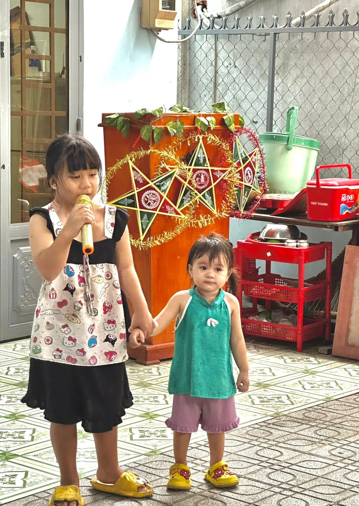 一个小女孩手持麦克风在大家面前讲话，身旁站着一个更小的孩子