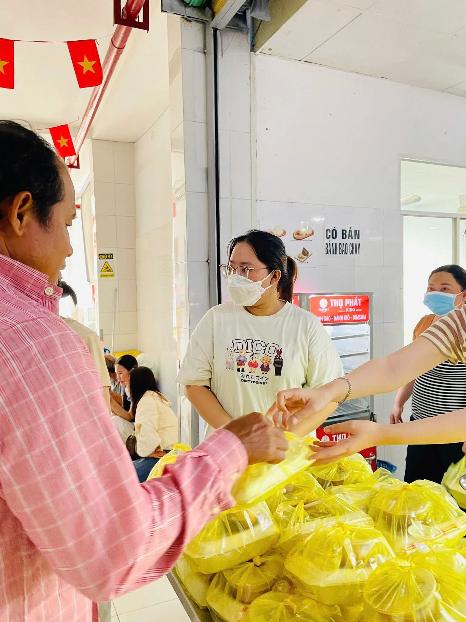 Tình nguyện viên trao phần ăn cho một người đàn ông mặc áo sơ mi caro hồng bên bàn đầy túi phần ăn màu vàng