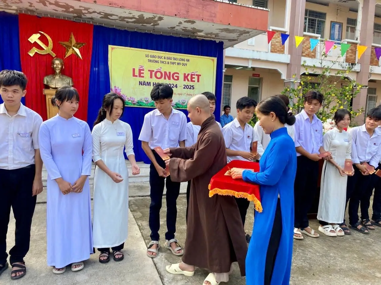 Một vị Thầy trao phong bì học bổng cho các em học sinh trước phông Lễ Tổng kết năm học 2024 – 2025 của Trường THCS & THPT Mỹ Quý