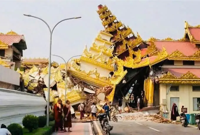 A multi-tiered golden-roofed temple building collapsed and leaning after the earthquake, with monks and local people standing on the street