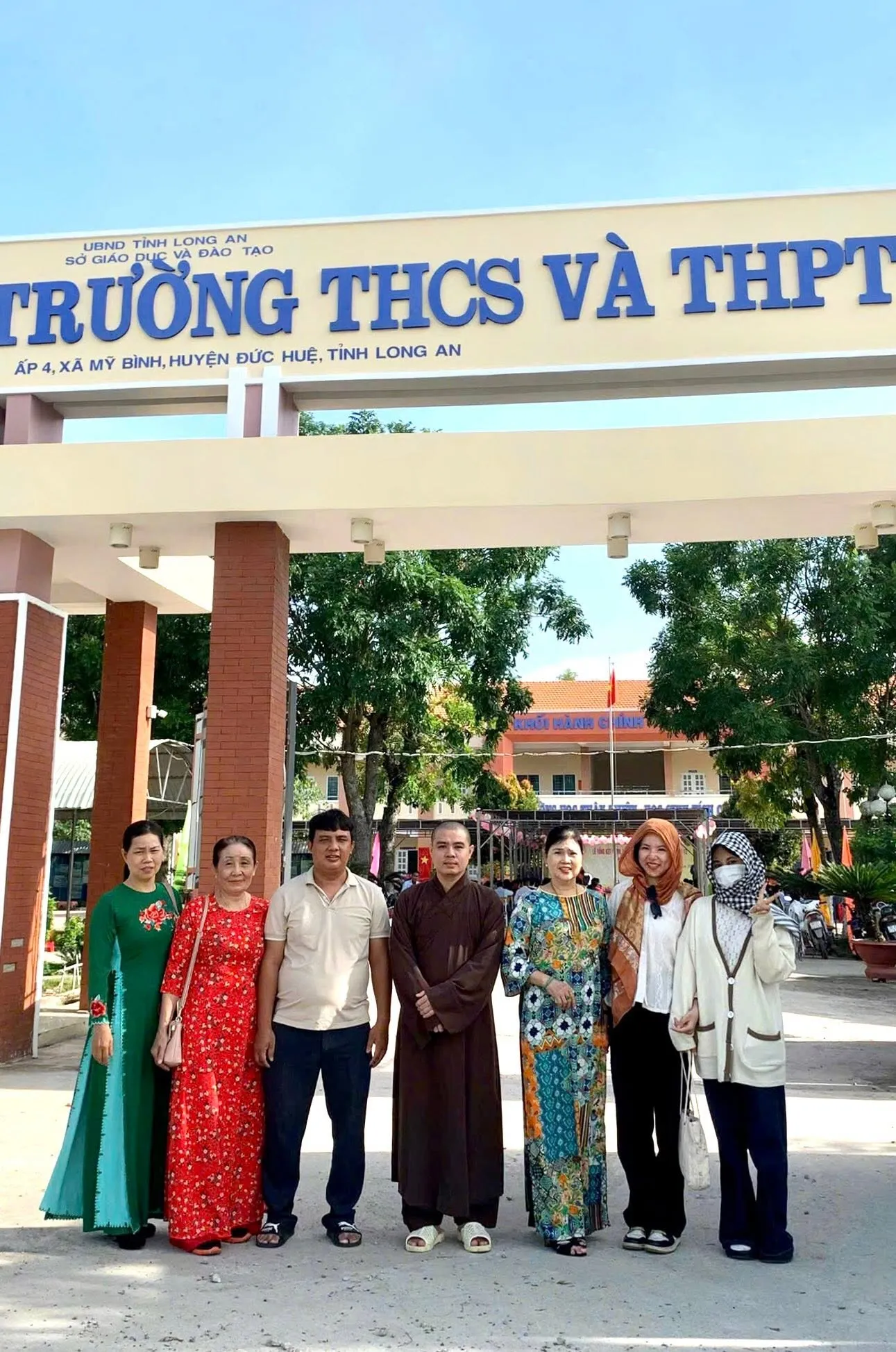 身穿褐色僧袍的法师与几位居士在学校（Trường THCS và THPT，Ấp 4, xã Mỹ Bình, huyện Đức Huệ, tỉnh Long An）校门前合影留念