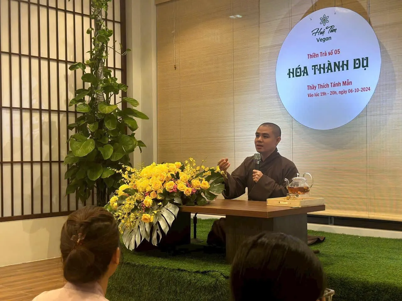 Một vị Thầy cầm micro chia sẻ bên bàn trà, cạnh bình hoa cúc vàng và tấm bảng Thiền Trà số 05 “Hóa Thành Dụ” tại Huệ Tâm Vegan