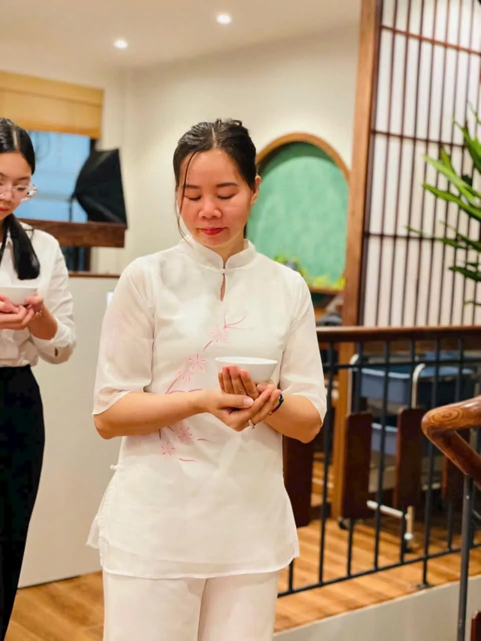Một người mặc áo trắng đứng, hai tay nâng chén trà, mắt nhìn xuống