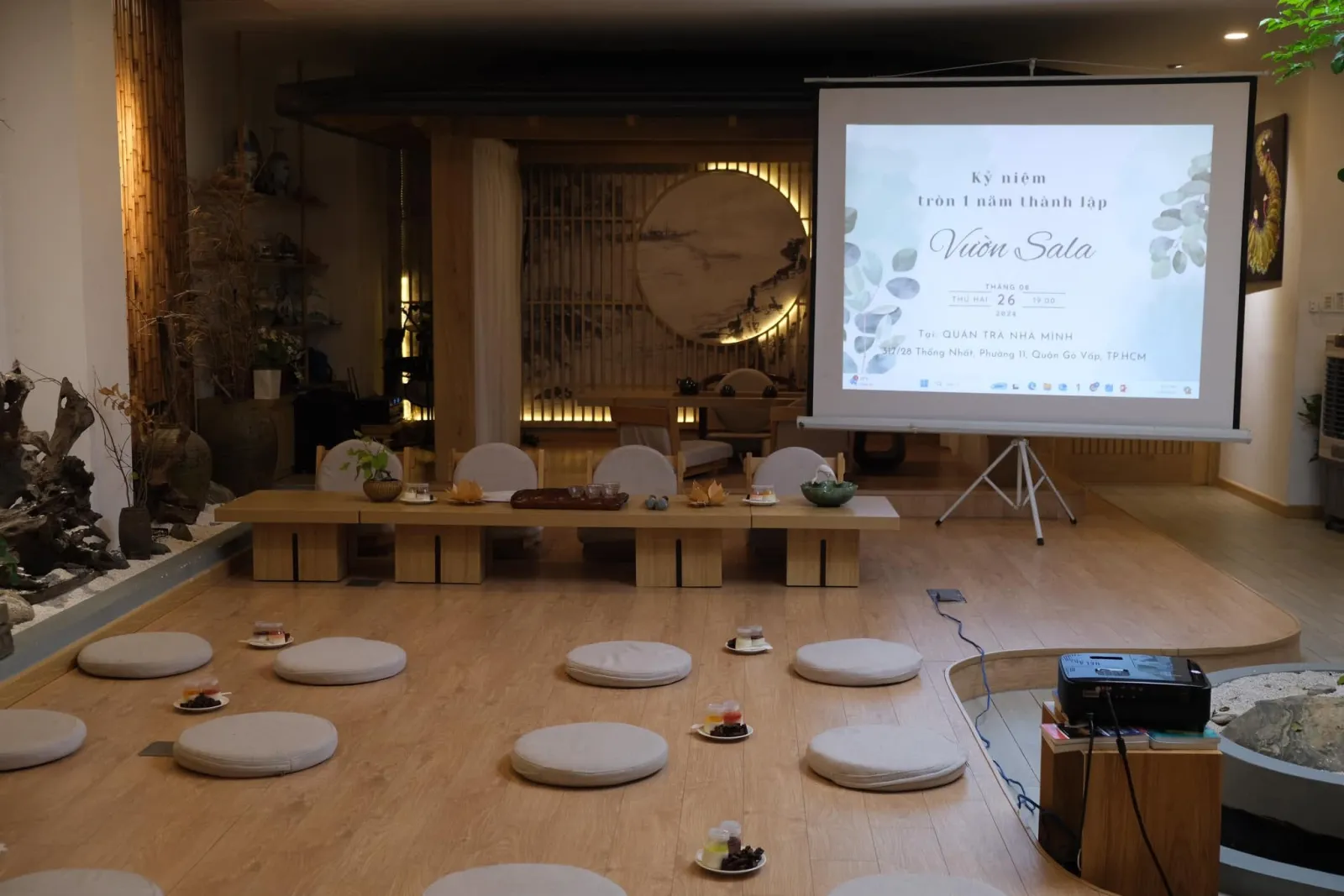 Projector screen reading “Kỷ niệm tròn 1 năm thành lập Vườn Sala” (1st anniversary of Vườn Sala), Monday 26 August 2024, 19:00, at Quán Trà Nhà Mình, with meditation cushions laid out in front