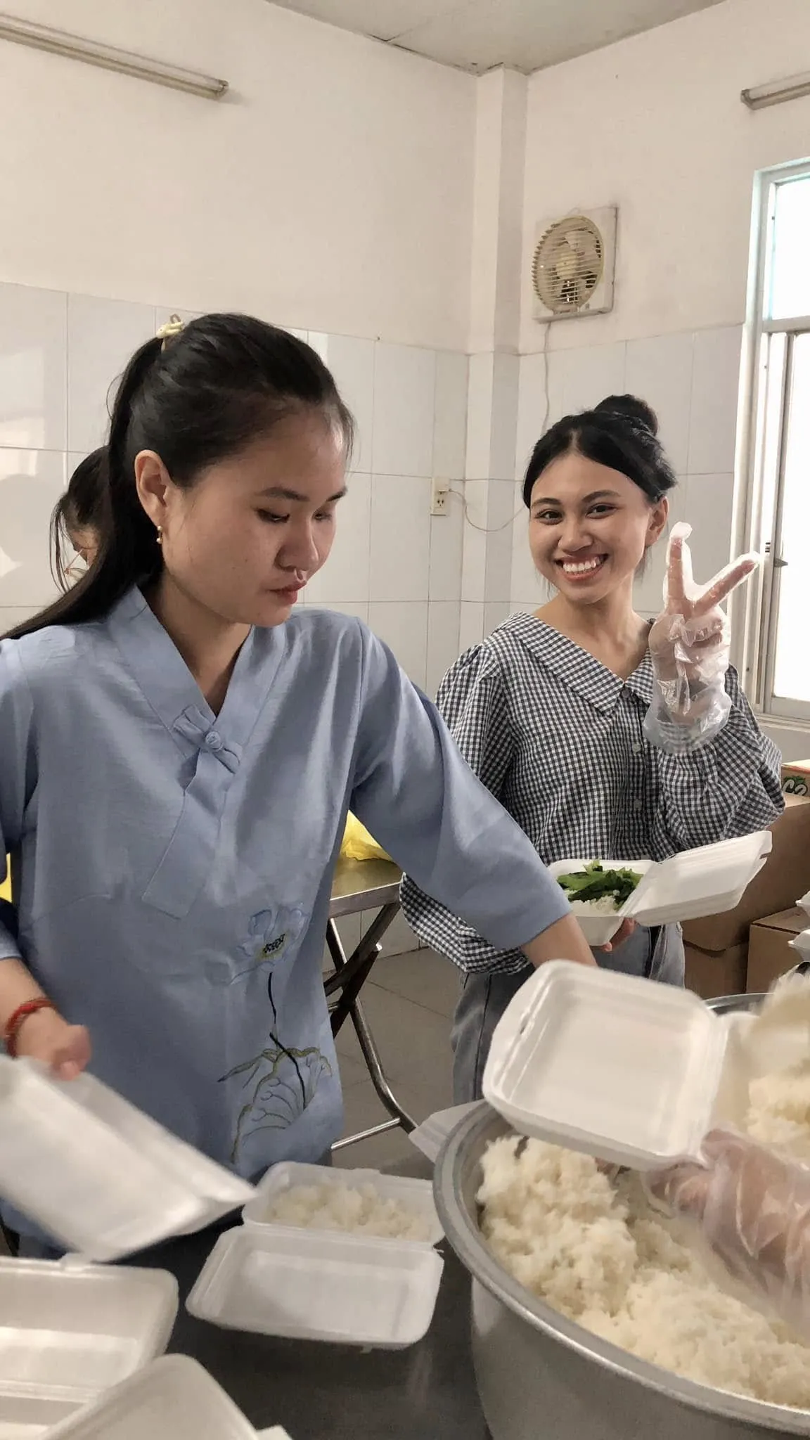 一位志愿者在一盆米饭旁拿着泡沫餐盒,另一位笑着比出V字手势