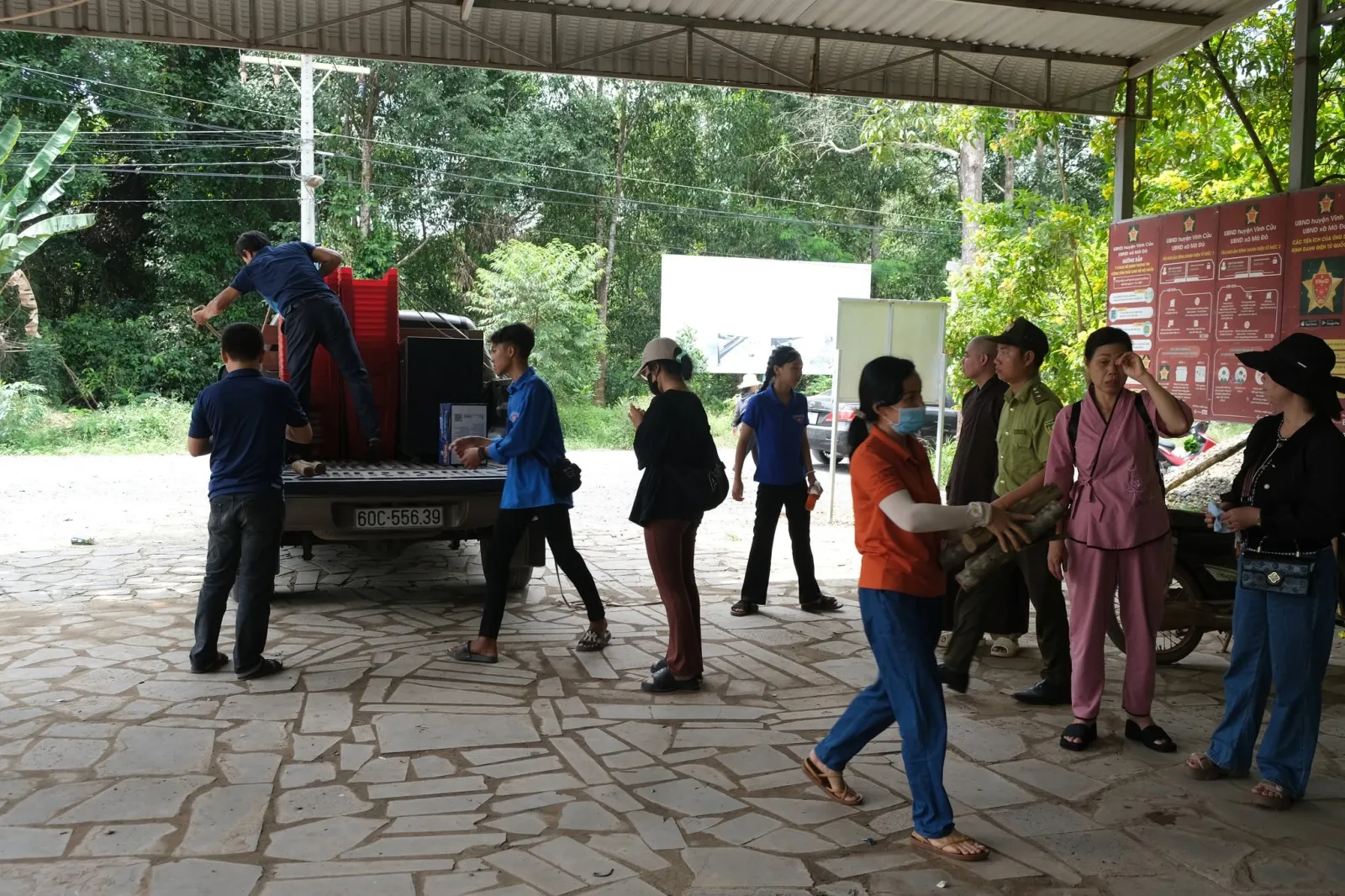 人们从皮卡车上搬运物品,旁边是 UBND huyện Vĩnh Cửu、UBND xã Mã Đà 的宣传栏