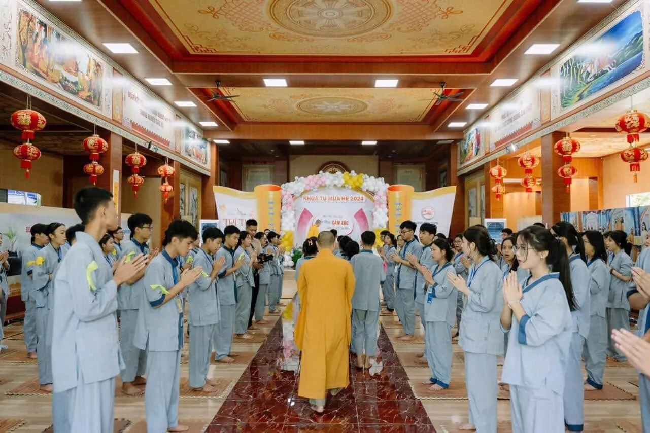 Retreatants in grey-blue uniforms stand with joined palms on both sides of the aisle as a monk walks into the hall towards the 2024 Summer Retreat stage