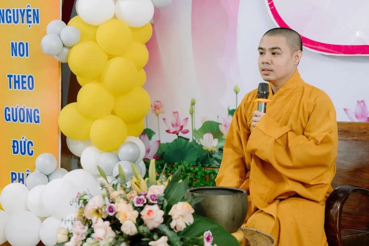 The Dharma teacher speaking into a microphone, seated beside flowers and a singing bowl