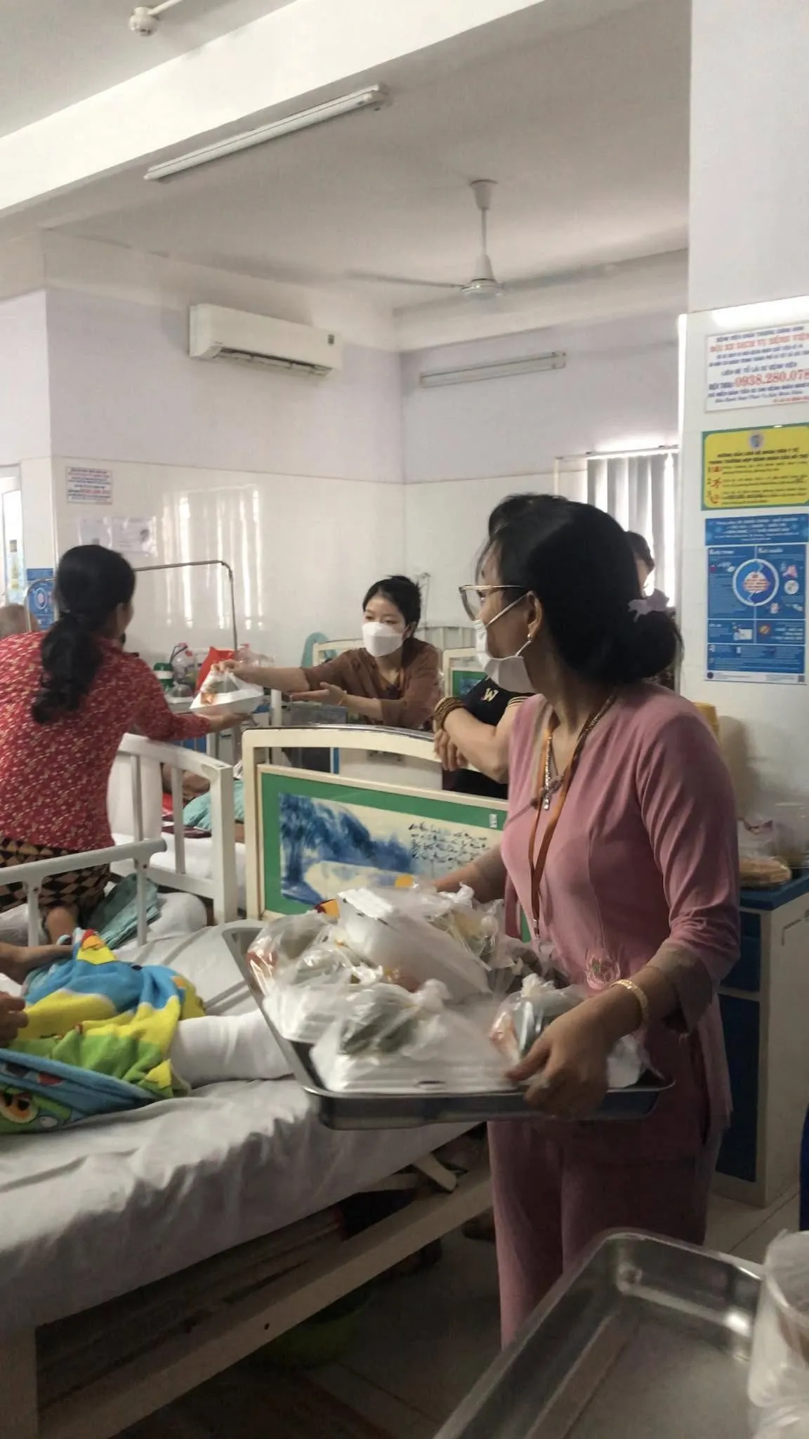 Một cô đeo khẩu trang bưng khay đầy túi phần ăn bên giường bệnh, phía sau một bạn tình nguyện viên trao phần ăn cho người bệnh
