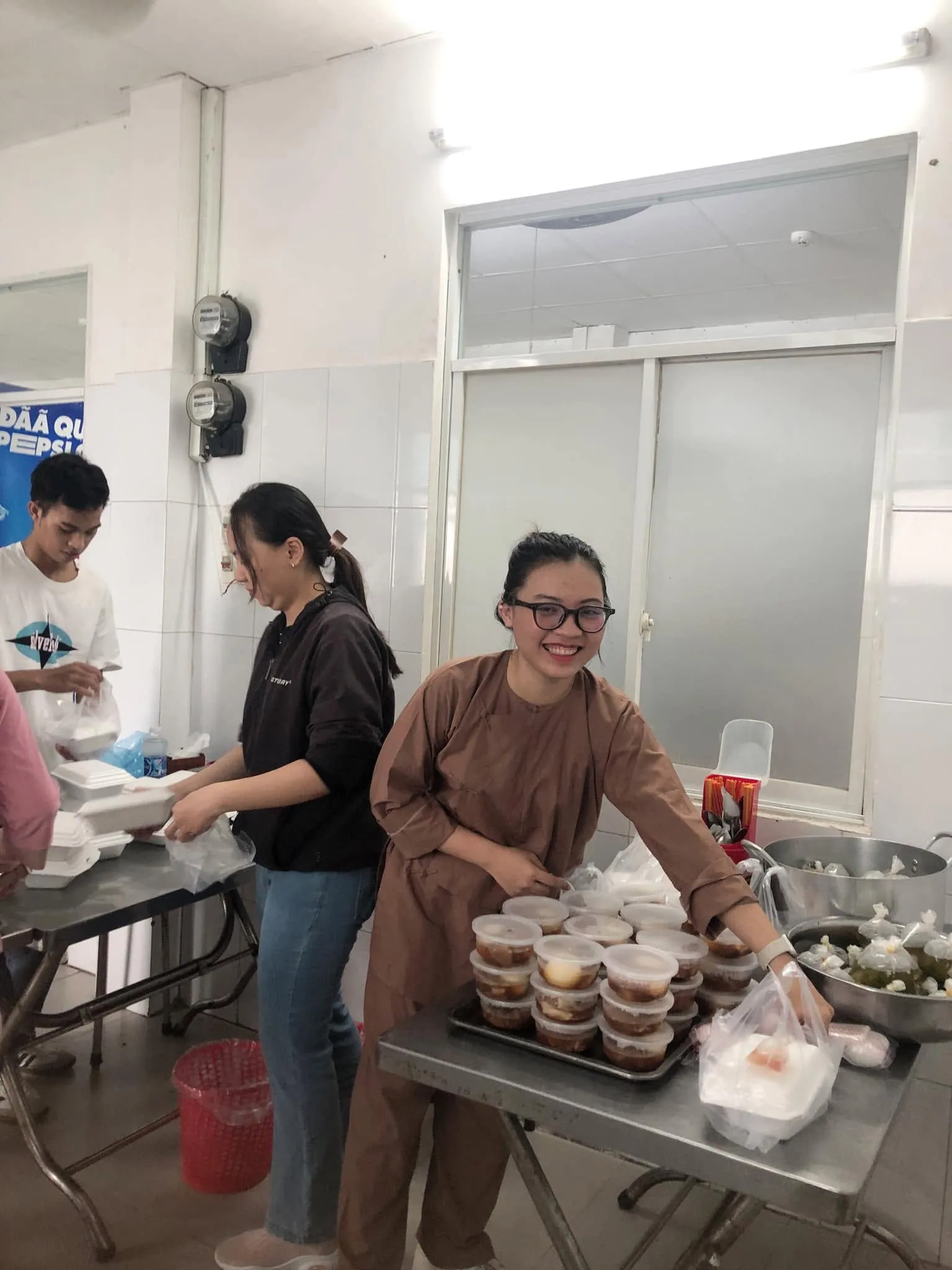志愿者们在打包餐食,一位身穿褐色衣服的志愿者站在一托盘带盖小餐盒旁微笑