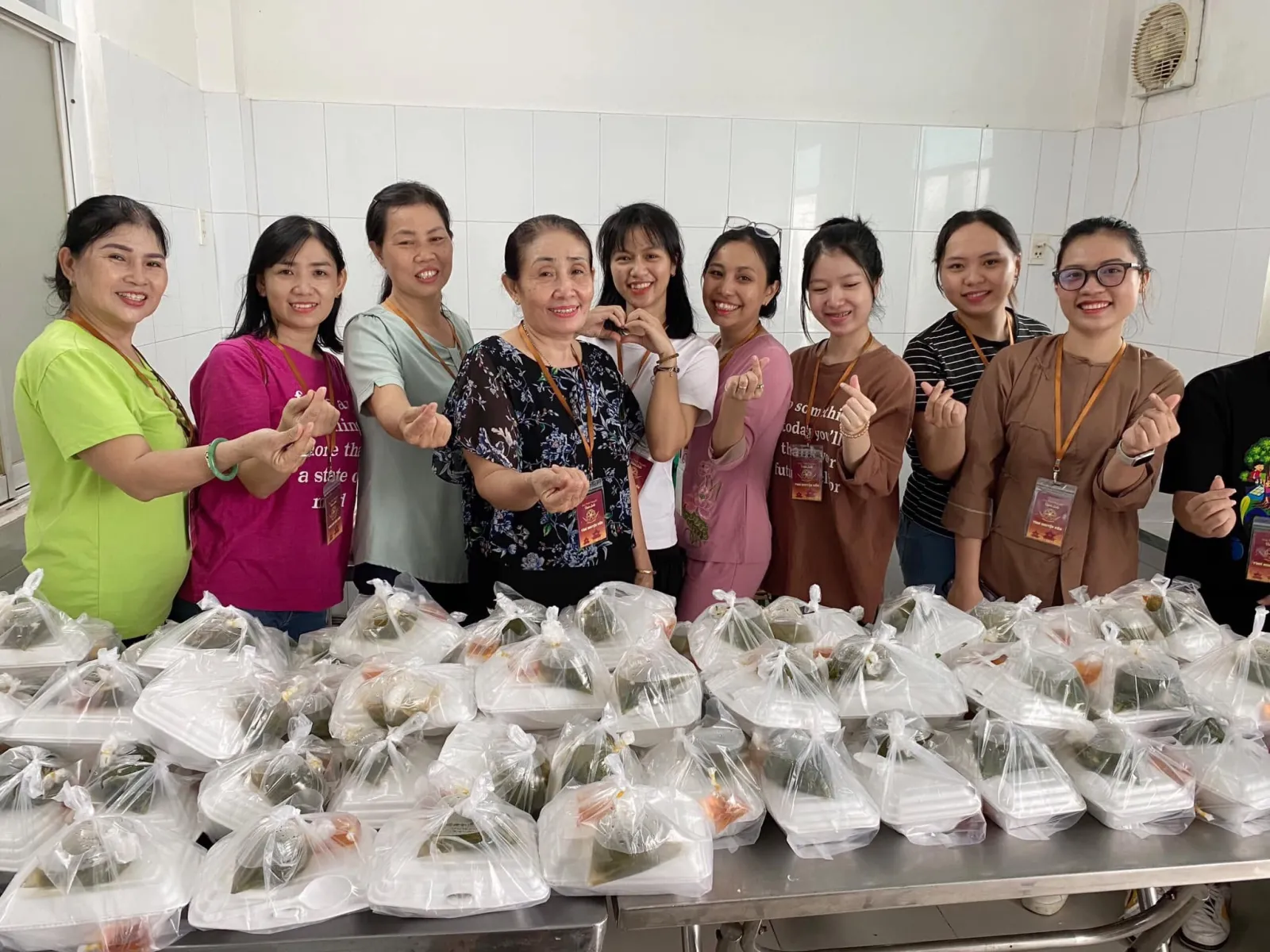 志愿者们站在桌上一排排餐袋后面,笑着比出手指爱心