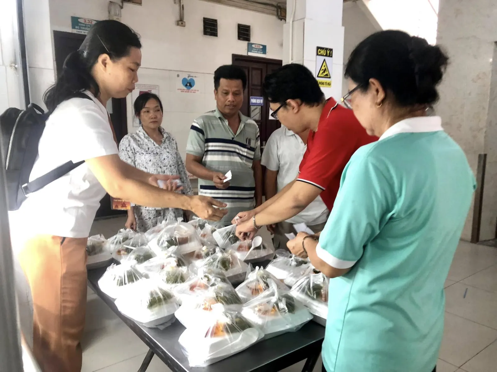 Các bạn tình nguyện viên trao phần ăn trưa cho bà con bên bàn đầy túi phần ăn trong hành lang bệnh viện