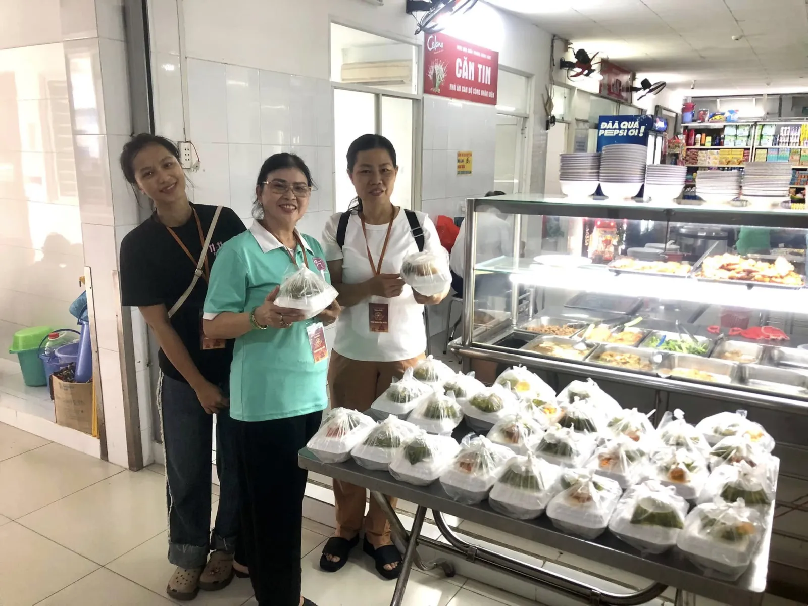 三位志愿者在医院食堂旁摆满餐袋的桌边,手捧餐食微笑