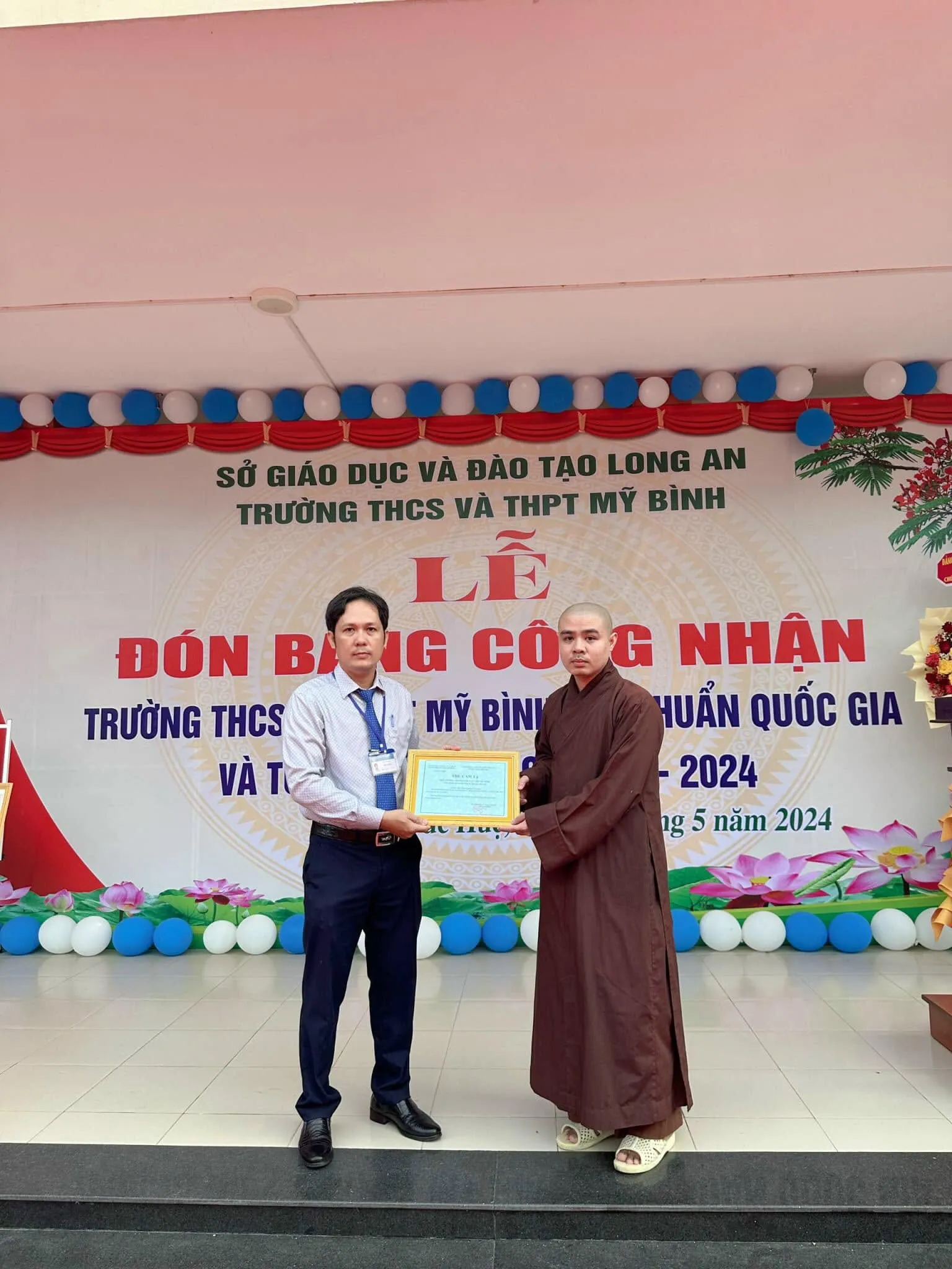 校方代表在 Trường THCS và THPT Mỹ Bình 典礼背景板前向身着褐色僧袍的法师递交感谢信