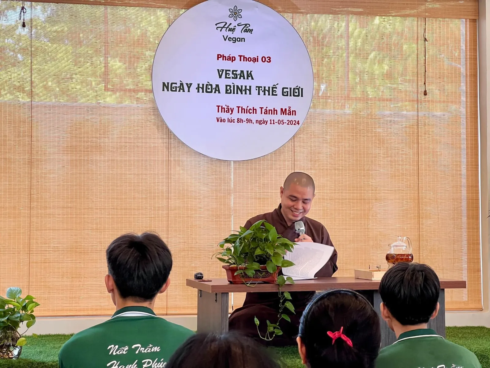 Thầy mỉm cười cầm micro và trang giấy bên bàn trà, người nghe ngồi phía trước