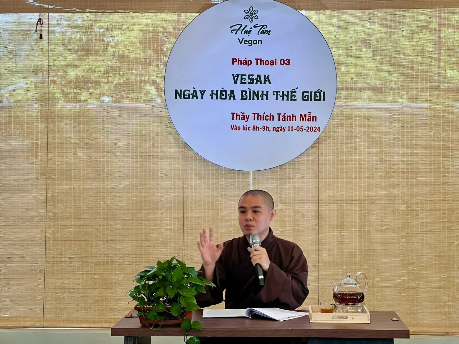 Thầy cầm micro giảng pháp bên bàn trà, phía sau là bảng tròn Pháp Thoại 03 “Vesak – Ngày hòa bình thế giới”